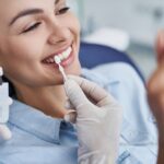 Woman smiling and sitting in a dental chair as a veneer is held up to compare to her teeth