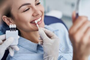 Woman smiling and sitting in a dental chair as a veneer is held up to compare to her teeth